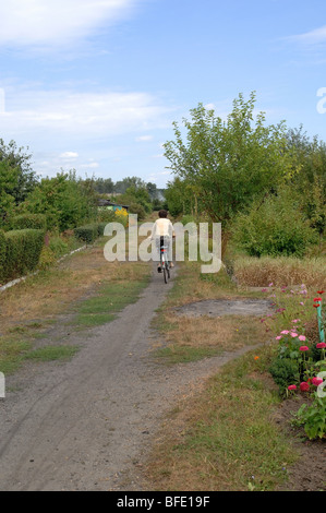 Uno di una serie di foto che ritrae una donna più anziana in bicicletta giù per un sentiero sterrato attraverso una sezione di singoli giardini. Foto Stock