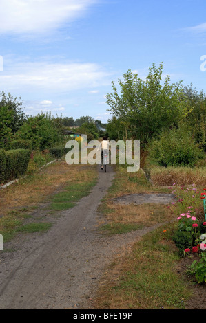 Uno di una serie di foto che ritrae una donna più anziana in bicicletta giù per un sentiero sterrato attraverso una sezione di singoli giardini. Foto Stock