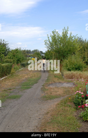 Uno di una serie di foto che ritrae una donna più anziana in bicicletta giù per un sentiero sterrato attraverso una sezione di singoli giardini. Foto Stock