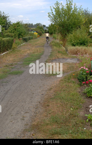 Uno di una serie di foto che ritrae una donna più anziana in bicicletta giù per un sentiero sterrato attraverso una sezione di singoli giardini. Foto Stock