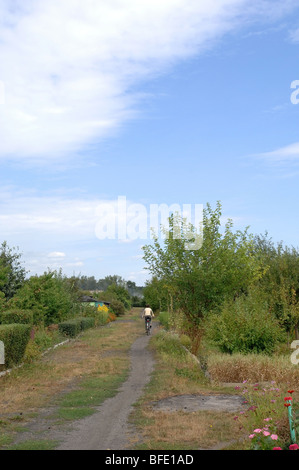 Uno di una serie di foto che ritrae una donna più anziana in bicicletta giù per un sentiero sterrato attraverso una sezione di singoli giardini. Foto Stock