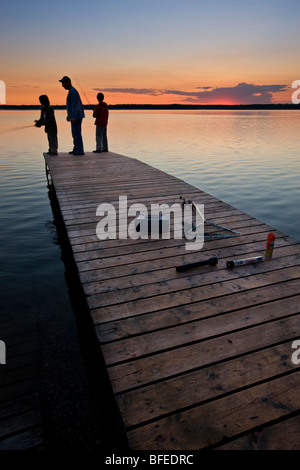 Un padre con i suoi due figli la pesca alla fine di un pontile sul Lake Audy al tramonto, Equitazione Mountain National Park, Manitoba, Canada Foto Stock