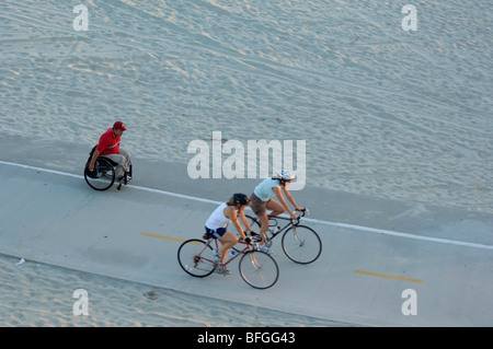 Due piloti di bicicletta il passaggio di una persona disabile che esercitano in una sedia a rotelle sulla stessa pista ciclabile. Foto Stock