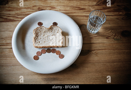 Bicchiere di acqua e il volto triste e realizzata in una fetta di pane raffermo e pochi centesimi su una piastra bianca Foto Stock