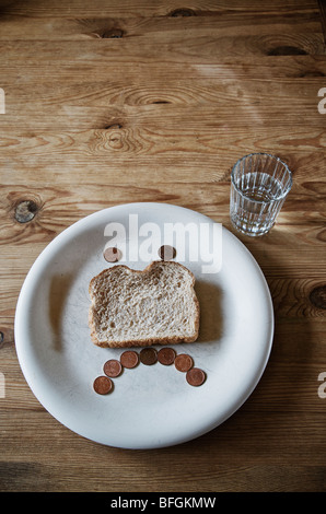 Bicchiere di acqua e il volto triste e realizzata in una fetta di pane raffermo e pochi centesimi su una piastra bianca Foto Stock