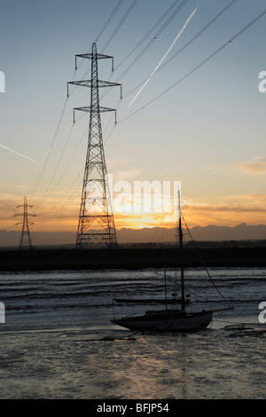 Tralicci di elettricità presso sunrise over Faversham creek nel Kent. Un piano lascia una contrail dietro di esso nel cielo. Foto Stock