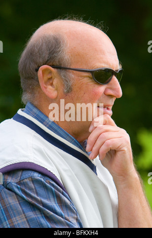 Un senior mentre maschio indossando occhiali da sole e tenendo la sua mento in piedi fuori, guardando lontano dalla telecamera e sorridente Foto Stock