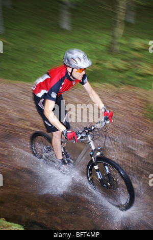 Scatto con sfocatura dinamica di un mountain biker che indossa un casco e schizza acqua mentre attraversa un ruscello nel bosco. Foto Stock