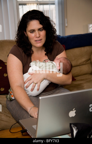 Nuova madre detiene il suo neonato mentre si lavora su un portatile Apple computer a casa nel suo soggiorno Foto Stock