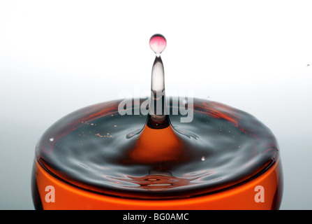 Una goccia di acqua rossa formando una colonna come esso cade in un bicchiere pieno di liquido, retroilluminato per il contrasto. Foto Stock