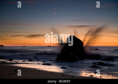 Un'onda si blocca su una grande roccia come una falce di luna tramonta sopra il Pacifico lungo la California's Big Sur Costa. Foto Stock