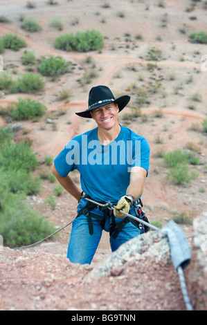 Una chiusura di un sorridente rocciatore prepararsi a rappel giù una montagna dopo una salita su un pendio ripido. Foto Stock