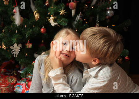 Ragazzo giovane dando la sua sorellina un bacio nella parte anteriore di un albero di natale con presenta qui di seguito Foto Stock