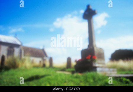 Impressionistica vista del sagrato in pietra memoriale di guerra con il crocifisso e il rosso dei gerani e erba con la chiesa sotto il cielo blu Foto Stock