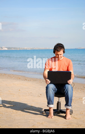 Giovane uomo seduto su una sedia in spiaggia a lavorare con un laptop Foto Stock