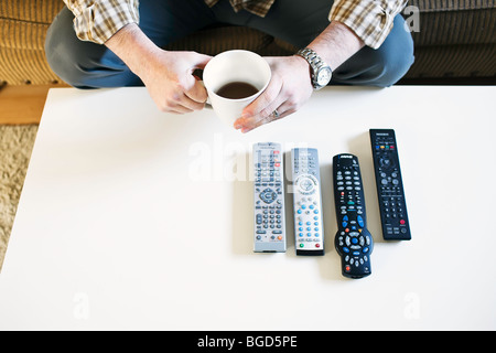 Uomo seduto sul lettino, tenendo il caffè, con telecomandi della TV schierate sul tavolo. Foto Stock