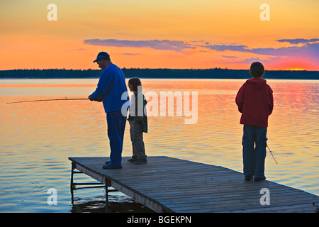 Un padre con i suoi due figli la pesca alla fine di un pontile sul Lake Audy al tramonto, Equitazione Mountain National Park, Manitoba, Canada Foto Stock
