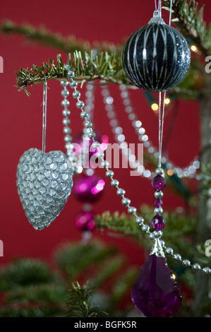 Un colorato a forma di cuore decorazione ninnolo splendidamente collocato su un pino albero di Natale su uno sfondo di colore rosso. Foto Stock