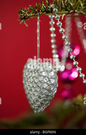 Un colorato a forma di cuore decorazione ninnolo splendidamente collocato su un pino albero di Natale su uno sfondo di colore rosso. Foto Stock