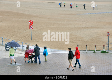 I turisti con ombrelloni camminando sul Seawall lungo la spiaggia in un giorno di pioggia durante le vacanze estive presso la costa belga Foto Stock