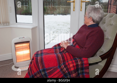 Anziani vecchia donna sola in casa con un tappeto sulle ginocchia mantenendo calda in ambienti chiusi da fuoco elettrico con la neve al di fuori delle finestre in inverno UK Gran Bretagna Foto Stock