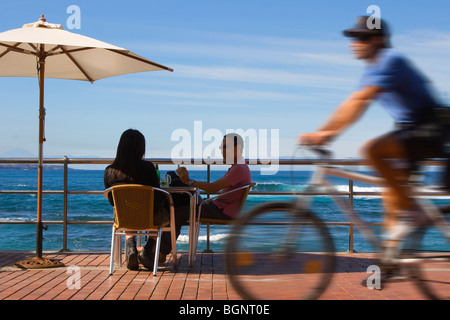 Giovane a un tavolo sulla spiaggia sotto un ombrellone con un ciclo di passaggio poliziotto Foto Stock