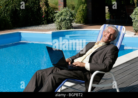 Happy businessman seduto sulla sedia a sdraio con computer portatile accanto alla piscina sorridente e guardando la fotocamera Foto Stock