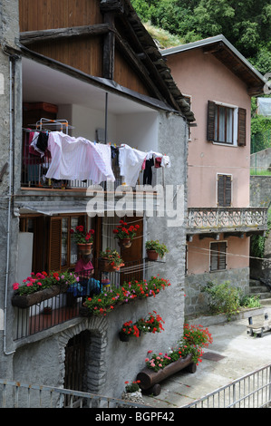 Old Lady cucito sul balcone circondato da finestre vasi per piante fiori rossi gerani rosa nella vecchia casa di Pont Saint Martin Aosta Foto Stock