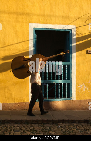 Un musicista che porta il suo contrabbasso passando accanto a una finestra di legno nella città coloniale di Trinidad de Cuba alla luce del sole serale Foto Stock