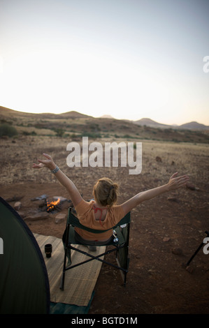 Vista posteriore della donna al tramonto al fuoco di campo con le braccia in aria, Sesfontein, Kaokoland, Namibia Foto Stock