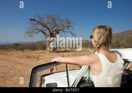 Giovane donna accanto al veicolo a motore guardando Baobab, Epupa Falls area, Kaokoland, Namibia Foto Stock