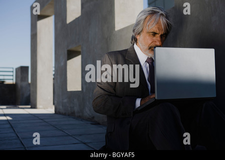 Uomo in tuta seduti sul marciapiede utilizzando laptop, edificio incompiuto in background Foto Stock