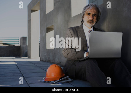 Uomo seduto sul marciapiede utilizzando laptop, elmetto impostato a lato, edificio incompiuto in background Foto Stock