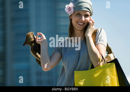 Donna con le borse della spesa parlando al cellulare, mantenendo la coppia di tacchi alti Foto Stock