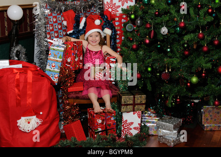 Poco ragazza seduta in Santa's chair circondato da regali di Natale e regali Foto Stock