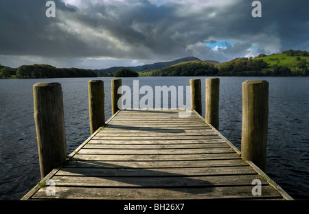 Una foto di un pontile raggiungendo in Coniston Water nel distretto del lago con drammatica nuvole e soleggiate colline basse. Foto Stock