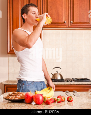 Giovane uomo che indossa il serbatoio superiore e jeans in cucina a bere un bicchiere di succo di arancia. Colpo verticale. Foto Stock