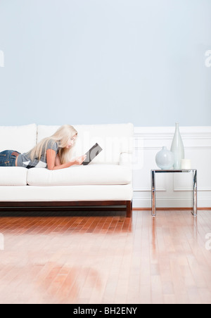 Giovane donna giace su un divano bianco in cima a scacchi e jeans blu durante la lettura di un libro. Colpo verticale. Foto Stock