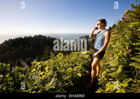 Una donna con occhiali da sole e vestiti estivi escursioni un sentiero costiero in Northwest Oregon e pause per rispondere il suo telefono cellulare. Foto Stock