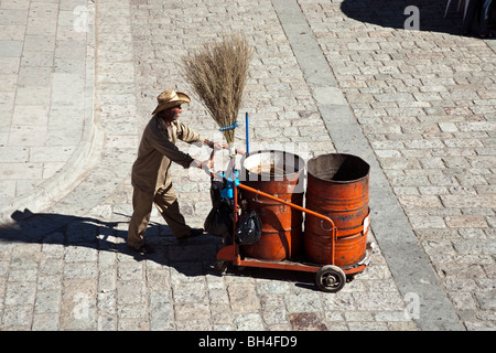 Messicano mustached street sweeper visto che attraversa un plaza nella città di Oaxaca spingendo il suo carrello con grandi frusta ginestra & Foto Stock