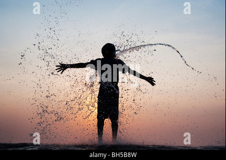 Ragazzo indiano avente acqua gettata su di lui contro un indiano al tramonto. Silhouette. Andhra Pradesh, India Foto Stock