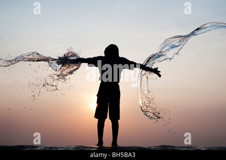 Ragazzo indiano avente acqua gettata su di lui contro un indiano al tramonto. Silhouette. Andhra Pradesh, India Foto Stock