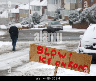 Cartello di avvertimento sul ghiaccio sulla strada suburbana e uomini in background clearing neve compattata. Foto Stock