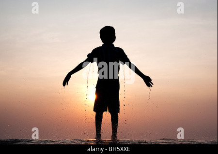 Ragazzo indiano gocciolante dopo aver acqua gettata su di lui contro un indiano al tramonto. Silhouette. Andhra Pradesh, India Foto Stock