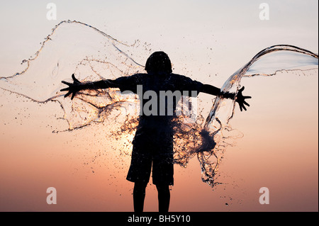 Ragazzo indiano avente acqua gettata su di lui contro un indiano al tramonto. Silhouette. Andhra Pradesh, India Foto Stock