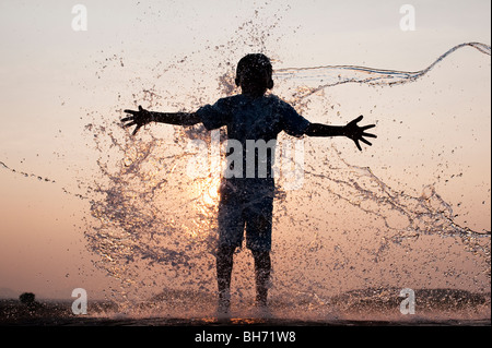 Ragazzo indiano avente acqua gettata su di lui contro un indiano al tramonto. Silhouette. Andhra Pradesh, India Foto Stock