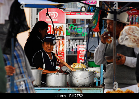 Ecuador, Otavalo, donna indigena di stare dietro a una pressione di stallo con patate saltate, profondo fritto di pollo e altri cotti soddisfare in bi Foto Stock