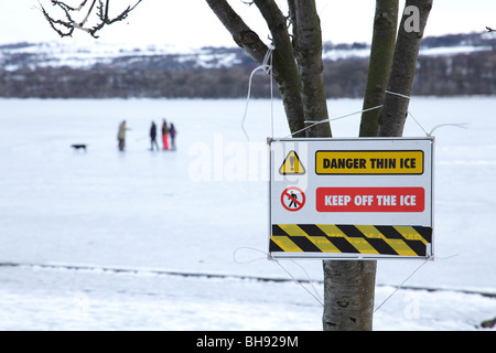 Il cartello di avvertimento di ghiaccio sottile accanto ad un Loch di Sapple del Castello congelato con la gente in inverno, il Parco Regionale di Clyde Muirshiel, Lochwinnoch, Renfrewshire, Scozia, Regno Unito Foto Stock