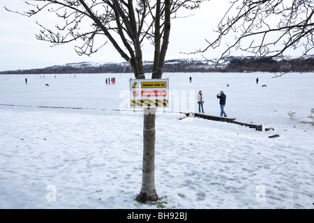 Il cartello di avvertimento di ghiaccio sottile accanto ad un Loch di Sapple del Castello congelato con la gente in inverno, il Parco Regionale di Clyde Muirshiel, Lochwinnoch, Renfrewshire, Scozia, Regno Unito Foto Stock