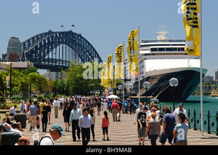 SYDNEY, Australia - turisti lungo il lungomare di Circular Quay di Sydney, con il Sydney Harbour Bridge sullo sfondo e una grande nave da crociera attraccata Foto Stock